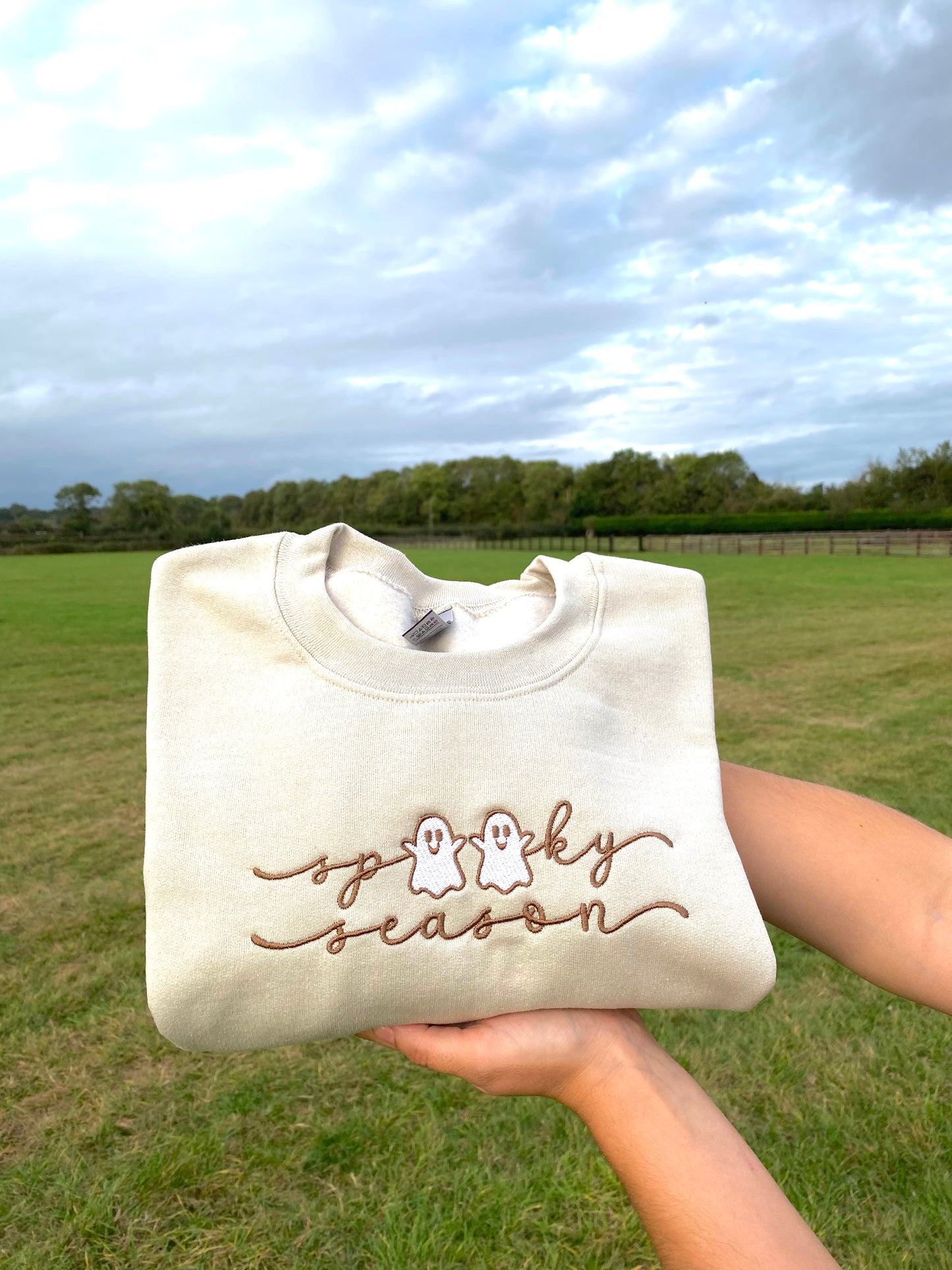 Beige sweatshirt with 'Spooky Season' embroidered text held in a field