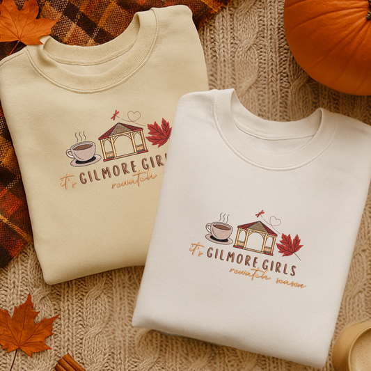 Two folded sweatshirts with 'Gilmore Girls' design on a textured surface with autumn leaves and a pumpkin.