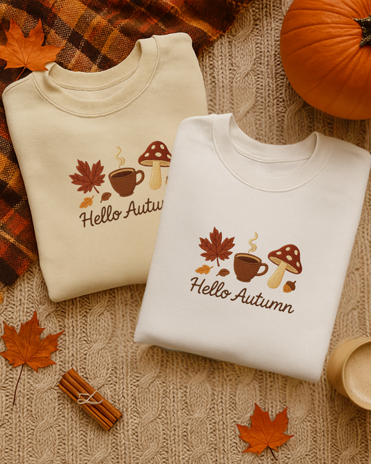 Two folded sweatshirts with an embroidered 'Hello Autumn' design on a textured surface with fall leaves and pumpkins.