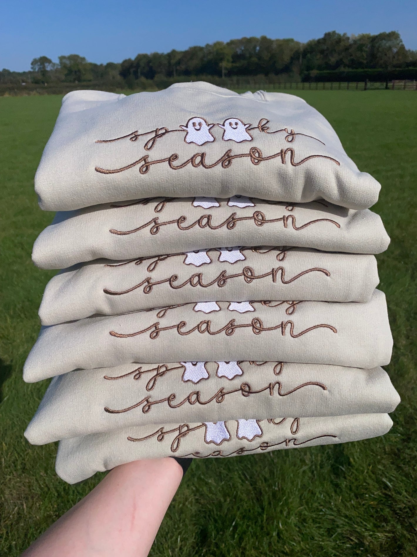 pile of folded cream sweatshirts with embroidered 'spooky season' ghost design