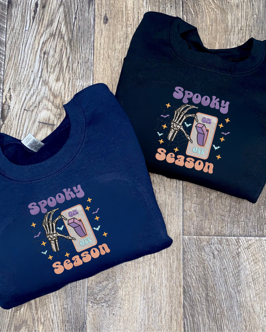a navy blue and a black sweatshirt with 'Spooky Season' embroidered design on a wooden surface